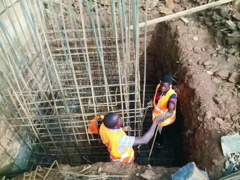 Two workers in safety vests setting a reinforcement cage in a foundation pit